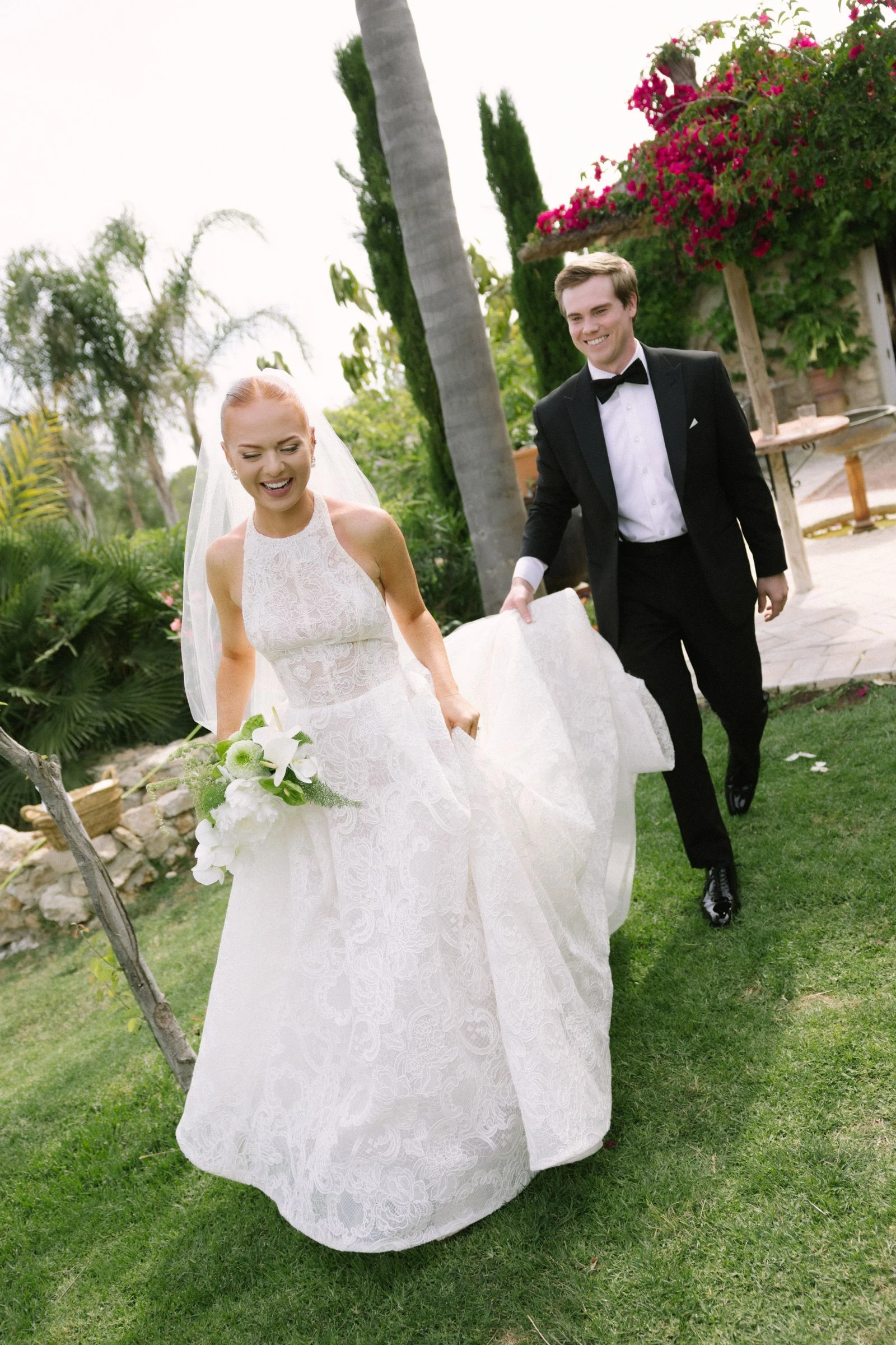 Novios caminando por el jardín, ella recogiéndose el vestido